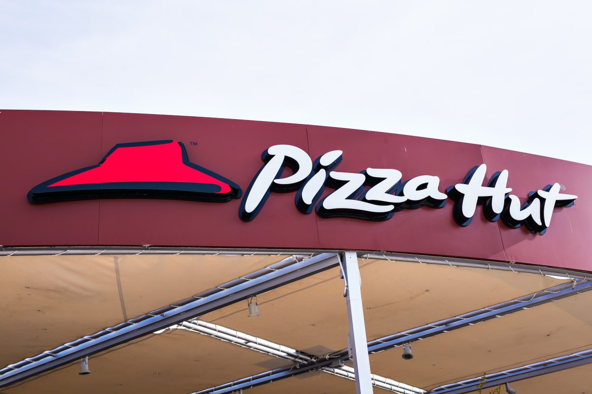 En röd skylt med den vita texten "Pizza Hut" med svart kontur och den tillhörande röda logotypen som liknar en hatt. Under skylten syns en ljus, beigefärgad markisliknande konstruktion med synliga metallramar och belysningsarmaturer.