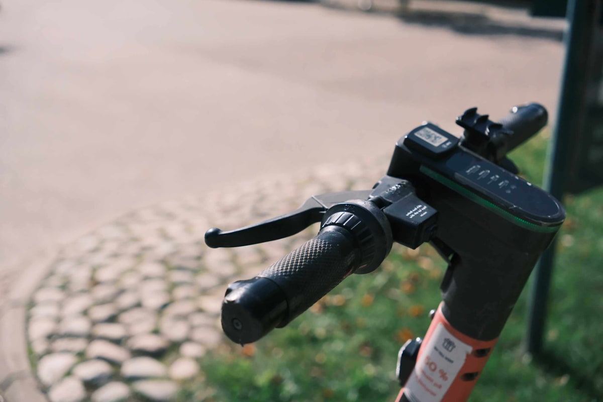 Bilden visar delar av styret och stången på en elsparkcykel i förgrunden, med ett handtag, bromshandtag och en display. I bakgrunden syns en kullerstensbelagd yta och gräs, delvis i höstfärger, under dagtid och utomhus.