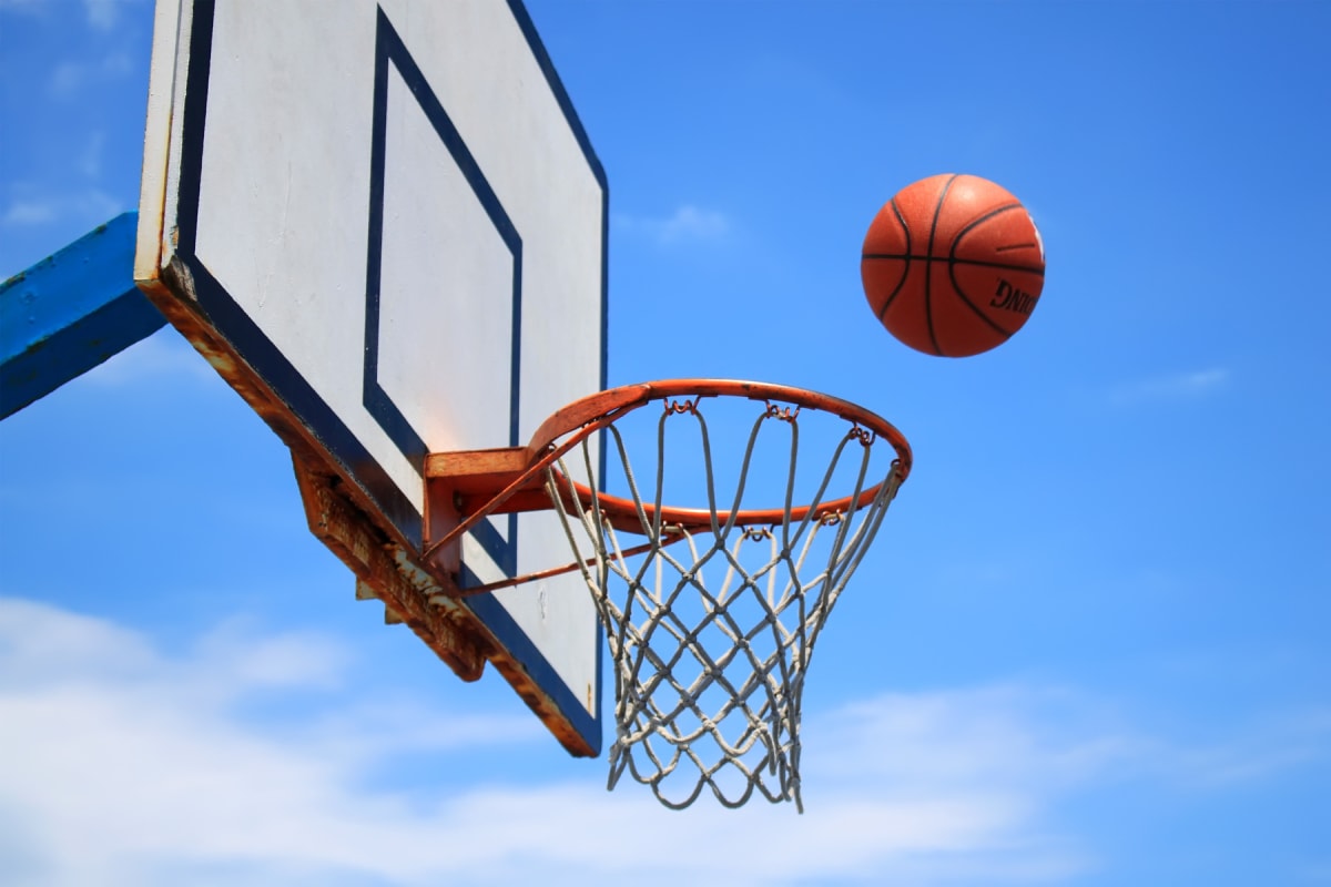 En orange basketboll är i luften nära en basketkorg med ett vitmålat bakstycke och en orange rostig ring. Bakgrunden består av en klarblå himmel med vita moln.