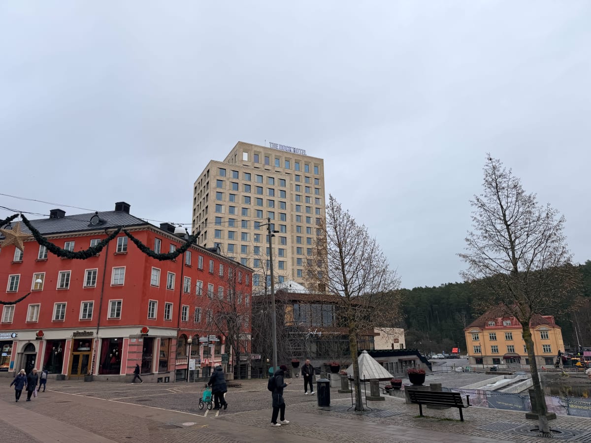 Bilden visar ett torg eller en öppen plats i en stad under dagtid. Till vänster syns en röd byggnad med vita fönster och juldekorationer i form av girlander och en stor stjärna. I nederkant av den röda byggnaden finns butiksfasader, bland annat en sk