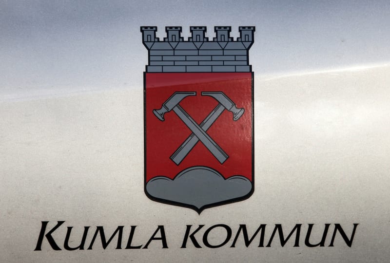 Bilden visar vapenskölden för Kumla kommun. Vapenskölden består av en röd sköld med två korsade silverfärgade hammare, symboliserande bergshantering eller industri. Nedanför hammarna finns ett silverfärgat moln. Ovanpå den röda skölden sitter en grå