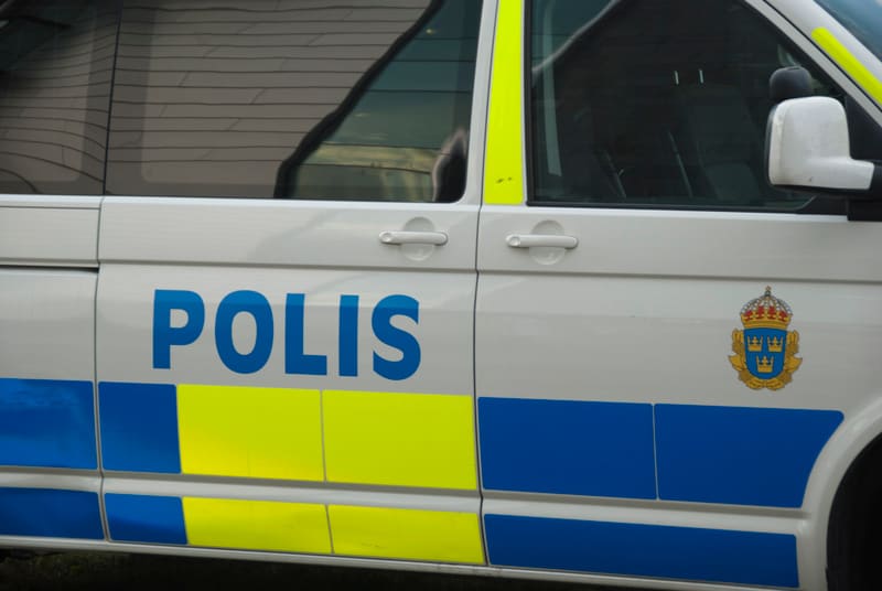 Bilden visar närbild på sidan av ett svenskt polisfordon. Fordonet är vitt med blå och neongula ränder samt texten "POLIS" i blå versaler. På bakre delen av fordonet syns Sveriges riksvapen i form av tre kronor och ett lejon, omgivet av ett gyllene b