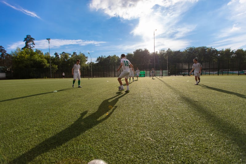 Bilden visar en grupp män som deltar i en fotbollsträning eller match på en konstgräsplan utomhus under dagtid. Flera spelare är klädda i vita tröjor och shorts, med vissa detaljer i grönt. En spelare i förgrunden med nummer 18 på ryggen dribblar en
