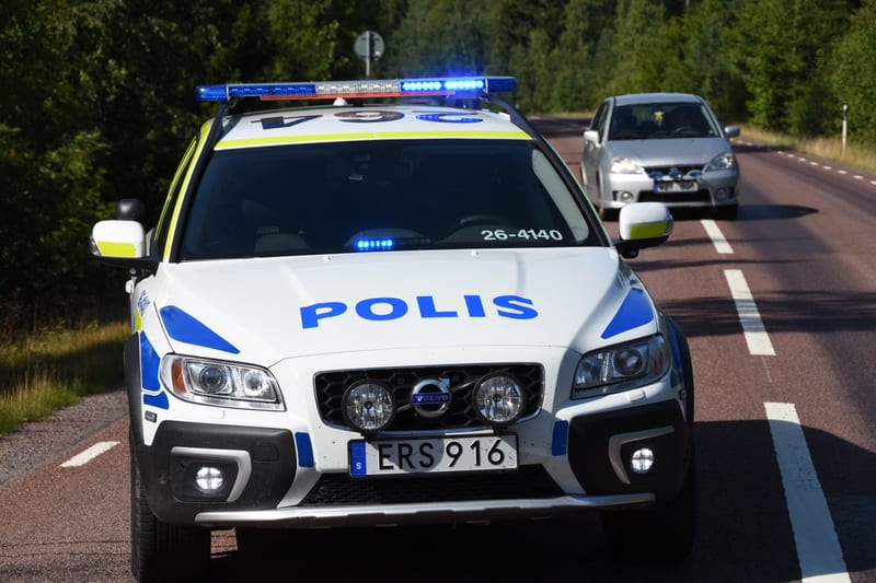En svensk polisbil av märket Volvo står på en asfalterad väg med skog i bakgrunden. Polisbilen har blått och gult mönster samt texten ”POLIS” på motorhuven. Blåljusen på taket och i fronten är tända. Bakom polisbilen, längre bort på vägen, syns en lj