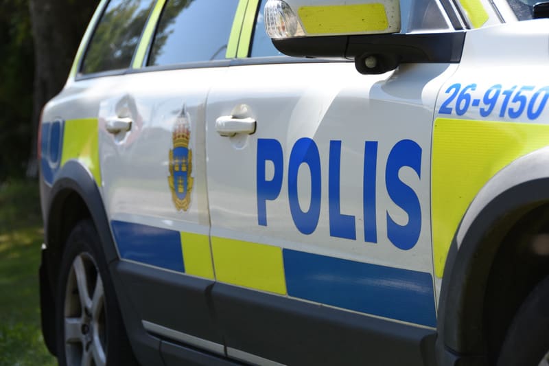Bilden visar närbild på sidan av en vit svensk polisbil. På bilens sida står ordet "POLIS" skrivet i stora blå versaler. Ovanför texten "POLIS" syns en del av polisens logotyp med lilla riksvapnet. Dörren har blåa och gula ränder som löper horisontel