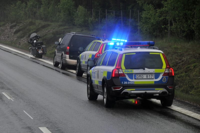 Bilden visar tre fordon parkerade längs sidan av en väg. Främst i bild står en svensk polisbil med blåa utryckningsljus aktiva. Bakom den står ytterligare en polisbil, delvis dold av den första. Längst bort, bakom den andra polisbilen och delvis uppe