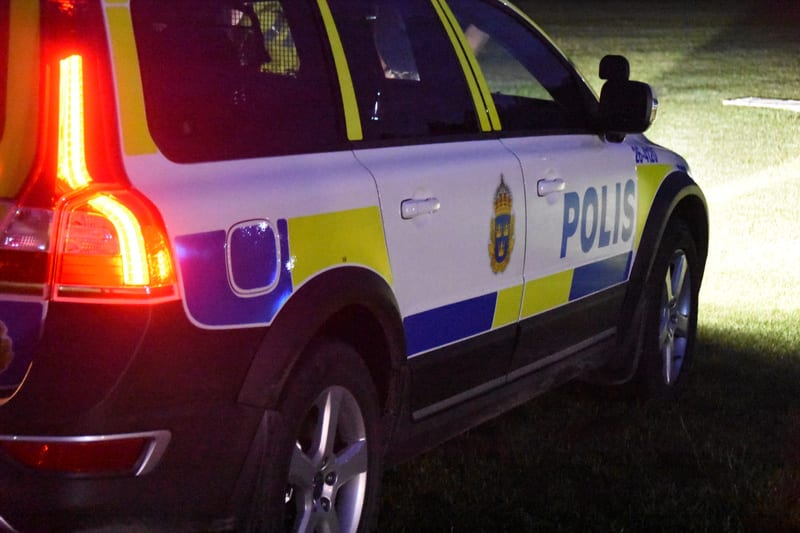 Bilden är en närbild av baksidan och sidan av en svensk polisbil av modellen Volvo. Baklyktorna på bilens vänstra sida lyser rött. På bilens sida framträder texten "POLIS" i blått med gul bakgrund, tillsammans med polisens emblem. Bilen verkar stå på