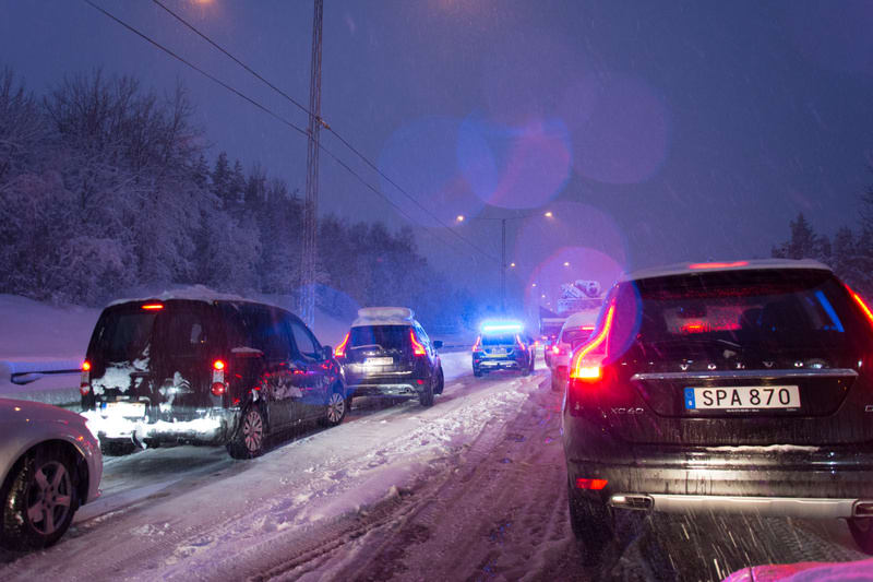 Bilden visar bilar i kö på en snötäckt väg under natten eller i skymningen. Vägen är täckt av snö och snöblandat slask. I bakgrunden syns träd täckta av snö, och det ser ut att snöa lätt. Flera baklyktor lyser röda och bromsljus lyser vitt. En bil lä