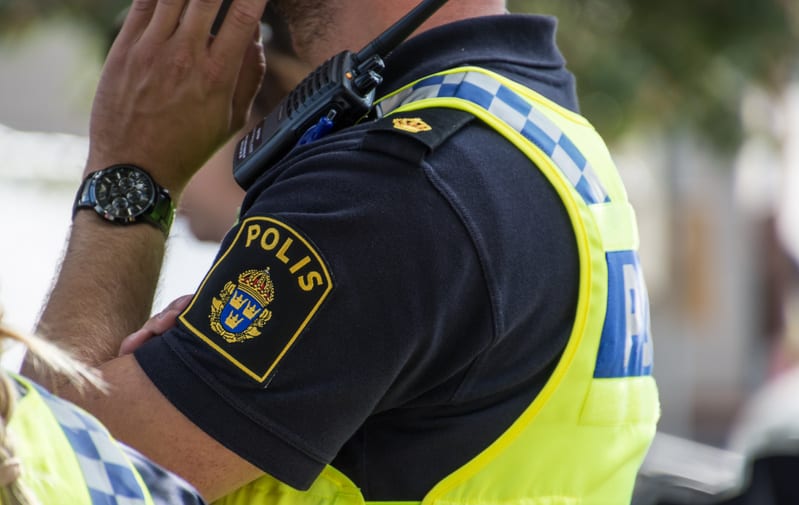 Bilden visar del av en persons överkropp, sannolikt en polis, med fokus på vänster arm och axel. Personen bär en mörkblå pikétröja med texten ”POLIS” och symbolen för Polismyndigheten (tre kronor över ett vapen) på en synlig patch på överarmen. Över