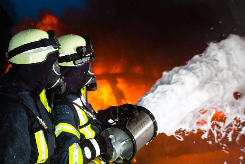 Bilden visar två brandmän i full skyddsutrustning, inklusive hjälmar, andningsmasker och skyddskläder med reflekterande detaljer. De befinner sig i en mörk miljö med en stor brandflamma i bakgrunden. En brandman håller en stor skumkanon som sprutar v