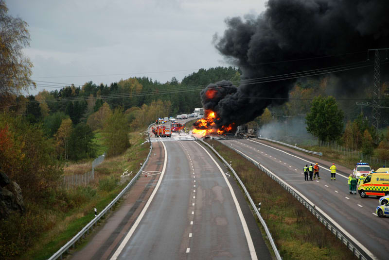 Bilden visar en motorväg med en pågående brand och räddningsinsatser. En stor svart rökpelare stiger upp från branden på vänster sida av motorvägen, där flera utryckningsfordon som brandbilar och ambulanser är placerade. Motorvägen ser ut att ha två