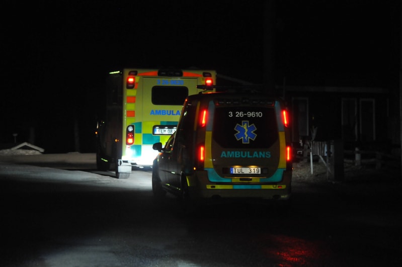 Bilden visar baksidan av två ambulansfordon i mörker. En större, gul och grön rutig ambulans med text 'AMBULANS' och 'ELM 081' syns i bakgrunden. Framför den står en mindre, gul- och blårandig bil med text 'AMBULANS', '3 26-9610' och en blå symbol so