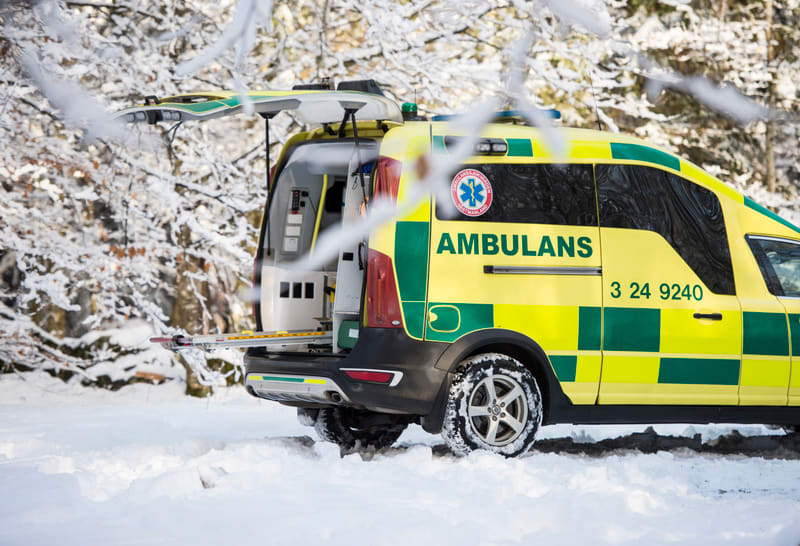 Bilden visar baksidan av en gul och grön ambulans av märket Volvo, som står i ett snötäckt landskap. Bakluckan på ambulansen är öppen, och en bår syns delvis dragen ut ur fordonet. Ambulansen har texten "AMBULANS" i stora gröna bokstäver på sidan, li