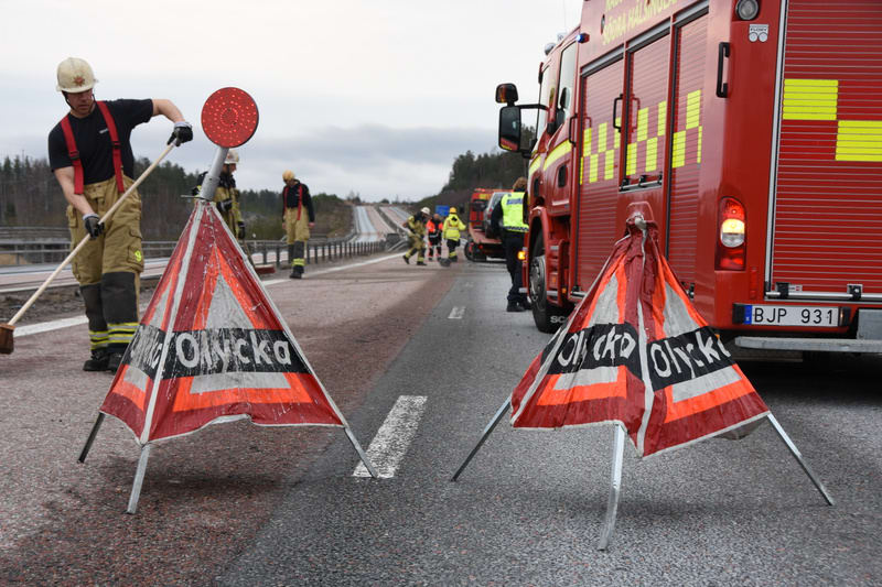 Bilden visar en vägsituation där räddningstjänst är på plats. I förgrunden syns två orangefärgade varningsskyltar med texten "Olycka" på, utplacerade på asfalten som ser ut att vara våt. Mellan skyltarna löper en bruten vit linje. En brandman i ljusf