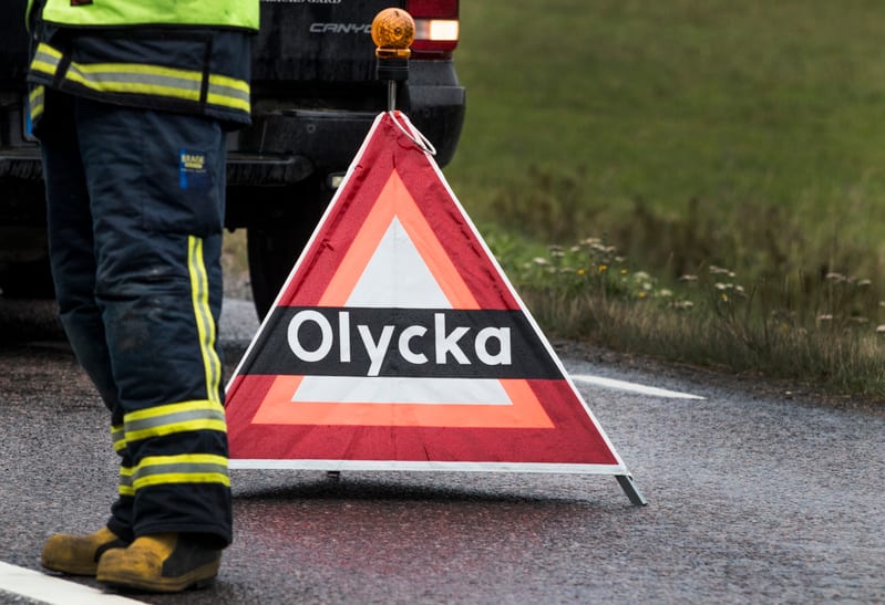 Bilden visar delar av en person, ett fordon och en varningstriangel på en våt asfaltsväg. Personen bär mörka byxor med gula reflexband och gula stövlar. En del av personens väst, som också har gula reflexer, syns. Bakom varningstriangeln syns en del