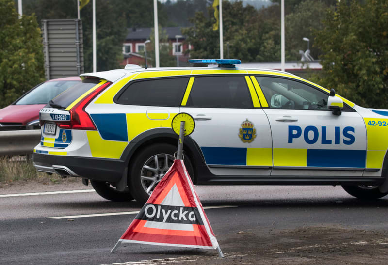 Bilden visar ett svenskt polisfordon, en vit kombi med blå och gul reflekterande dekor, samt texten "POLIS" och Polismyndighetens emblem. Fordonet står på en vägbana. På taket av polisfordonet syns en blå ljusramp. Framför polisfordonet, på vägbanan,