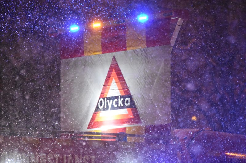 Bilden visar ett mörkt vinterlandskap under natten med snöfall. I förgrunden syns delar av ett fordon, troligtvis en utryckningsbil, med blinkande blå och orange varningsljus. På fordonets bakdel eller en påhängd skylt sitter en stor varningstriangel