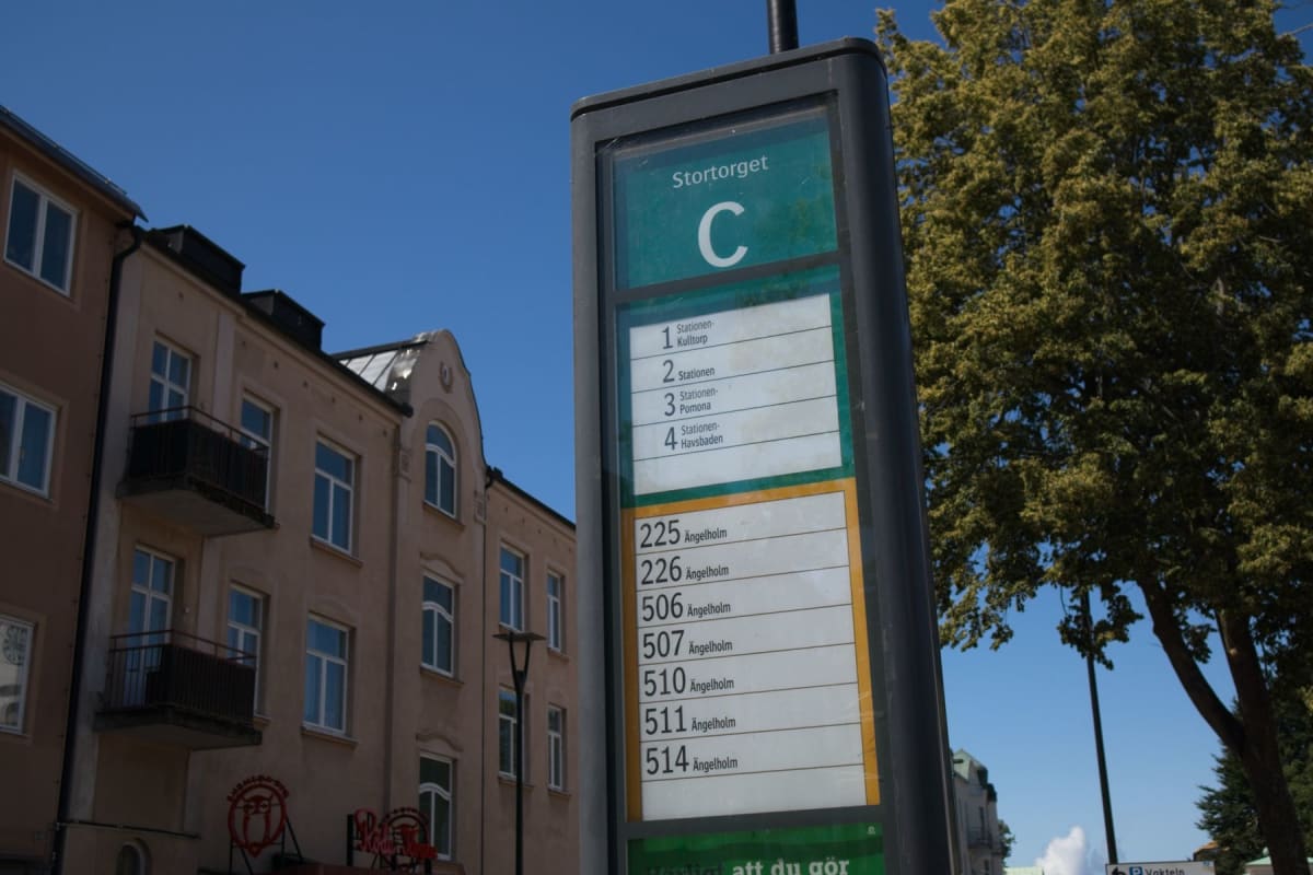 Bilden visar en busskylt vid en hållplats som heter 'Stortorget C'. Skylten listar busslinjer med nummer och destinationer som 'Ängelholm' samt stationer som 'Kultorp', 'Pomona' och 'Havsbaden'. I bakgrunden syns en flervåningsbyggnad med ljusrosa fa