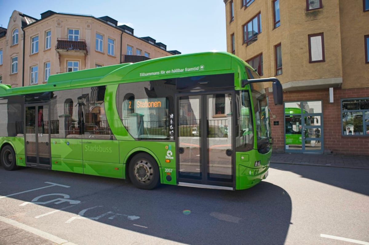 En ljusgrön stadsbuss, märkt 'Stadsbuss' och 'Skånetrafiken', står parkerad på en gata i dagsljus. Bussen har texten 'Tillsammans för en hållbar framtid' på taket och destinationsskylten visar '2 Stationen'. Hjul och bussdörrar är synliga. I bakgrund