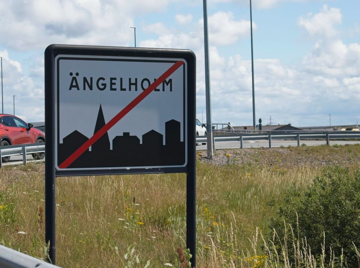 Bilden visar en skylt märkt "ÄNGELHOLM" med ett rött diagonalt streck över. Under texten finns en svart siluett av en stadssiluett med en kyrkspira. Skylten står vid sidan av en väg som är synlig i bakgrunden. På vägen syns en röd bil och en vit bil.
