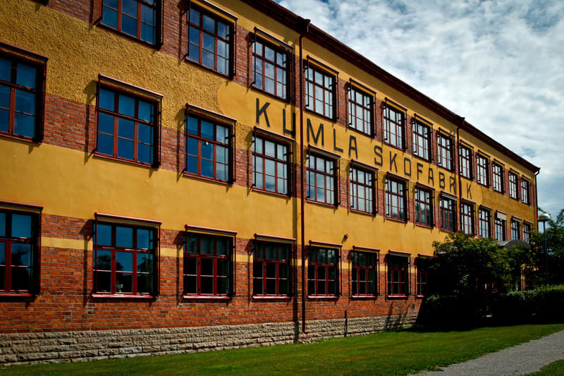 Bilden visar fasaden på en gul byggnad med många fönster. Fönstren har röda karmar och reflekterar himlen. Byggnadens nedre del är av rött tegel och under det finns en grå stenfot. Mellan tegelraderna och den gula väggen finns en horisontell röd linj