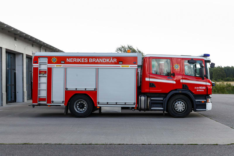 En röd brandbil med vita och silverfärgade detaljer står parkerad på en asfalterad yta framför en byggnad. Brandbilen har texten "NERIKES BRANDKÅR" i vitt på sidan och ett emblem som liknar en gul stjärna eller sol. Vid hytten finns också nummer och