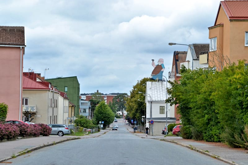 Bilden visar en gata i en stadsmiljö under dagtid. Gatans mitt är fri från fordon men parkerade bilar syns längs sidorna. Byggnader i olika färger som ljusrosa, beige och grå med röda tak omger gatan. På ett vitt hus i bildens högra del ses en stor f