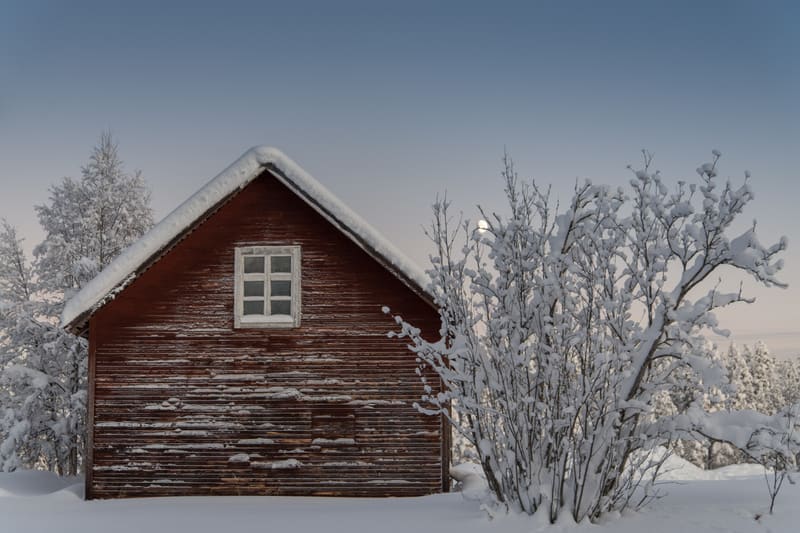 Bilden visar en ensam röd trälada med ett vitt fönster mitt på gaveln. Taket och marken runt ladan är täckta av ett tjockt lager snö. På ladans vänstra sida syns snötäckta träd och buskar, och på höger sida en större, snötyngd buske. Bakom busken och