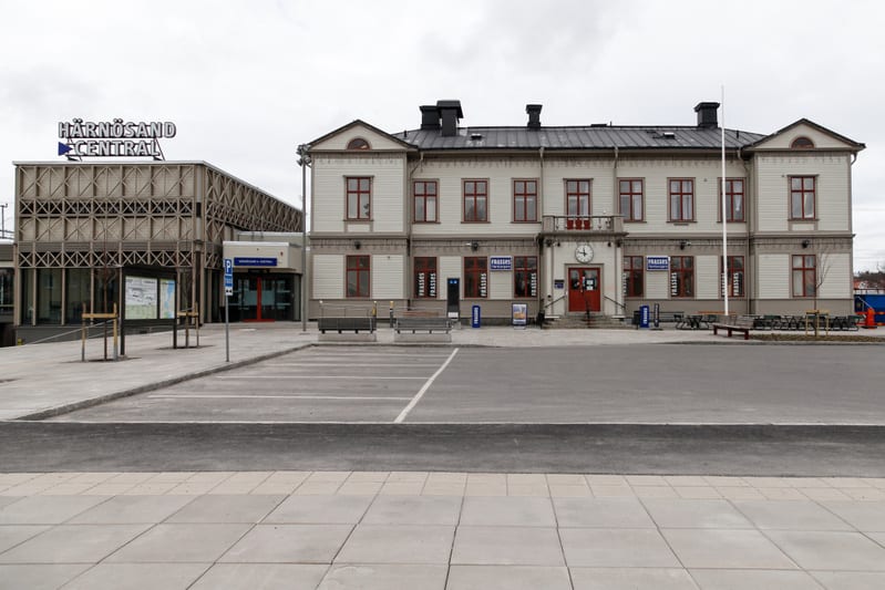Bilden visar Härnösands centralstation under en mulen dag. Framför stationen finns en parkeringsplats med markerade rutor och en gångväg. Stationen består av två delar: en äldre byggnad i ljus färg med rött tak och rödbruna fönsterramar, samt en mode