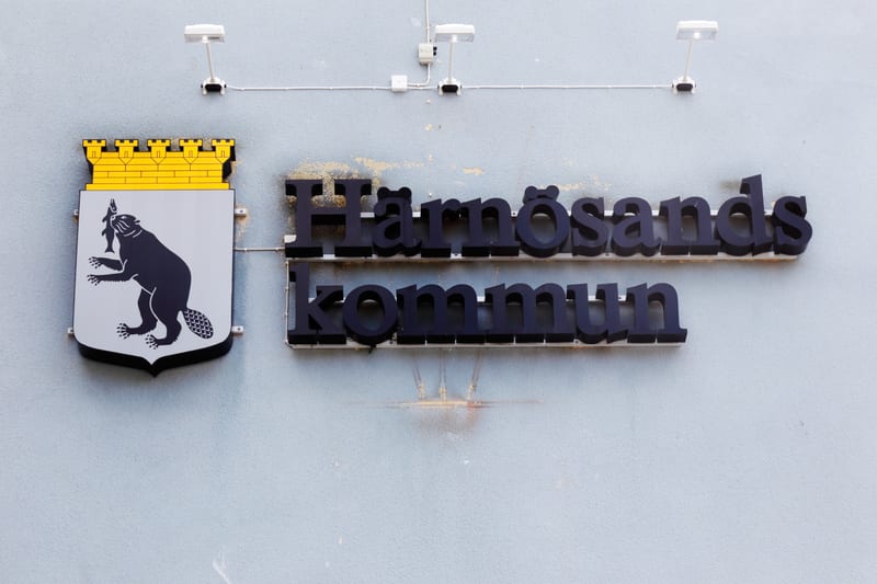 Bilden visar vapenskölden för Härnösands kommun tillsammans med texten "Härnösands kommun" i svarta, upphöjda bokstäver på en ljusblå vägg. Vapenskölden är vit med en stiliserad svart bäver som håller en fisk i tassarna och en guldfärgad murkrona ova