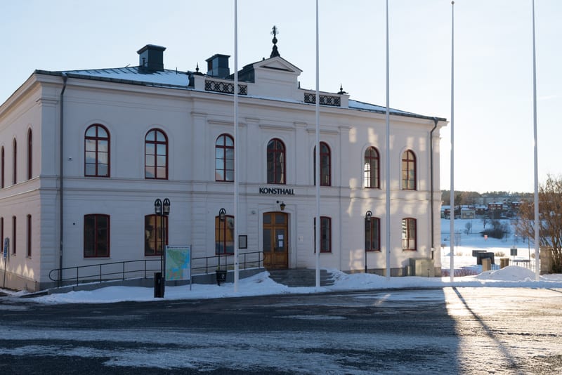 En stor, vit byggnad, sannolikt en konsthall baserat på texten ovanför entrén, står i fokus i en vinterlandskapsbild. Byggnaden har många fönster med bågar och en utsmyckad taklinje. Framför entrén finns en trappa samt en ramp för tillgänglighet, omg