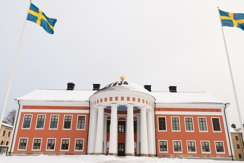 Bilden visar en stor byggnad i fokus, sedd rakt framifrån. Byggnaden är målad i en varm röd/orange färg med vita detaljer ovanför och under fönstren, samt runt en central entré. Den centrala entrén har en rundad form med pelare och ett kupolformat ta