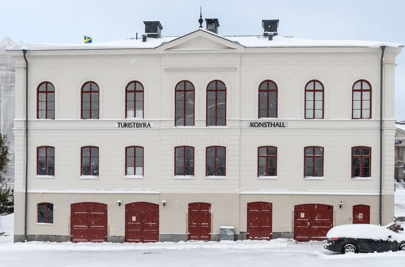 En fasadbild av en gräddvit, tvåvåningsbyggnad med en svensk flagga på taket. Byggnaden har flera brunröda fönster med välvda överdelar på övervåningen och rektangulära fönster på nedervåningen. Mellan våningarna finns skyltar med texten "TURISTBYRÅ"