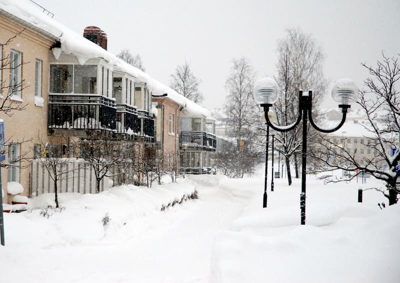 Bilden visar ett snötäckt bostadsområde under vintertid. Flera bostadshus med ljusrosa fasader och balkonger, varav några är inglasade, kantar en snötäckt gångväg. Taken på husen är täckta med tjocka lager av snö med istappar hängande från takfoten.