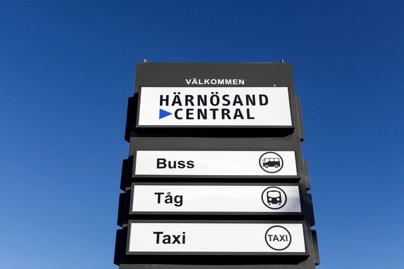Bilden visar en skylt för Härnösand Central, placerad utomhus mot en klarblå himmel. Skylten består av flera paneler. Överst står 'VÄLKOMMEN' och direkt under 'HÄRNÖSAND CENTRAL' med en blå pil pekande åt höger. Nedföljande paneler indikerar 'Buss',