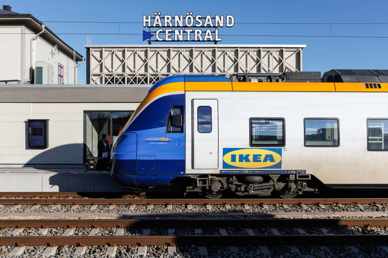 Bilden visar ett tåg, Norrtåg, parkerat vid en perrong. Tåget har blå, gul och vit färgsättning med IKEA-logotypen framträdande på sidan. I bakgrunden syns en byggnad med en skylt som säger "HÄRNÖSAND CENTRAL" med en pil som pekar åt vänster. En del