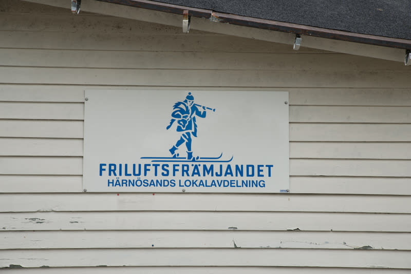 Bilden visar en vit rektangulär skylt med blå text och en blå logotyp monterad på en ljus, horisontell träfasad. Fasaden har synliga sprickor och flagnande färg. Ovanför skylten, i bildens övre del, syns en del av ett mörkgrått tak med ett rostfärgat