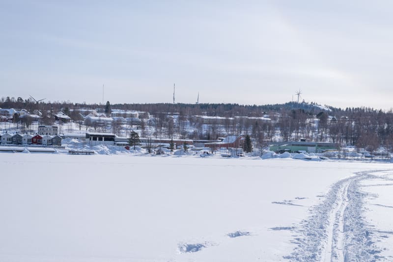 En landskapsbild tagen under dagtid med en bred horisontell vy över ett snötäckt landskap. I förgrunden syns ett vidsträckt område täckt av snö, troligen en frusen sjö eller ett fält, med tydliga skidspår som löper diagonalt från höger sida och inåt