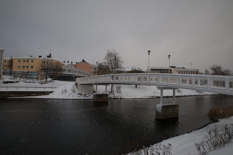 Panoramabild som visar ett snötäckt stadslandskap under en gråmulen himmel. I förgrunden flyter en mörk vattenyta, förmodligen en å eller flod, och längs strandkanterna ligger snö. En vit gångbro med räcken korsar vattnet och leder till den bebyggda