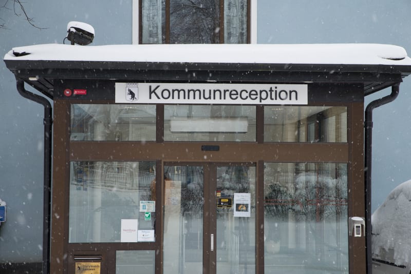 En bild tagen utifrån som visar entrén till en byggnad med skylten "Kommunreception" ovanför dörrarna. Det är snöigt och snöflingor syns i luften. Taket ovanför entrén är täckt med snö och det syns även en del snö på marken till höger om dörrarna. Dö
