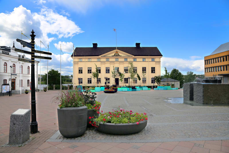 Bilden visar ett torg med flera byggnader under en delvis molnig blå himmel. I förgrunden syns stora grå blomkrukor med olika växter och blommor. Torget är belagt med gatsten i olika nyanser. Till vänster i bilden står en svart skyltstolpe med flera