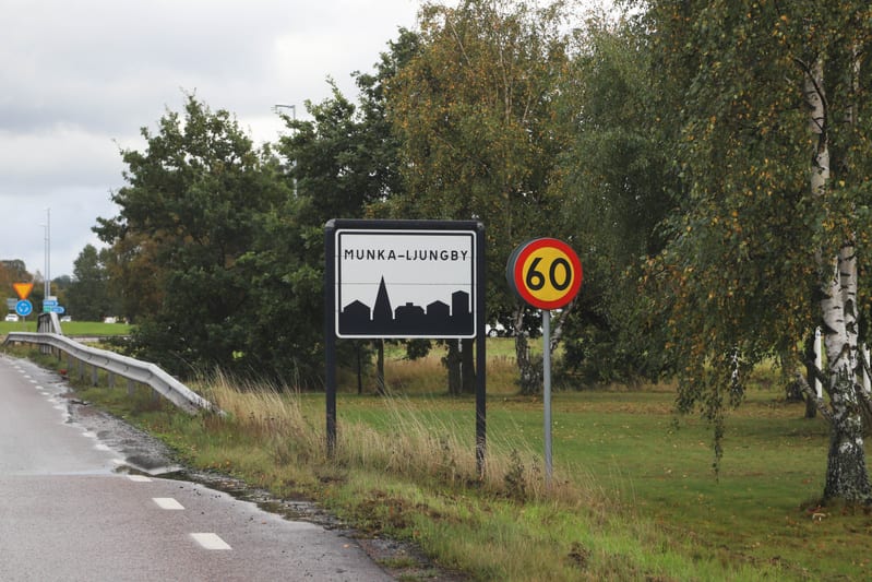 Bilden visar en utomhusmiljö med en asfalterad väg i förgrunden som har vita streckmarkeringar. Till höger om vägen finns en gräsbevuxen dikeskant följt av ett område med tätare och högre gräs. Där står två skyltar: en rektangulär skylt med texten "M