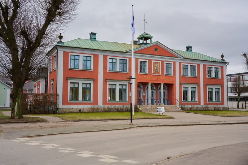 Bilden visar ett stort, rektangulärt, laxrött tingshus med vita detaljer och ett grönt tak. Framför byggnaden finns en gräsmatta och en gångväg. Byggnaden har många fönster med gröna fönsterkarmar och en markant entré med pelare och flera dörrar. Ova
