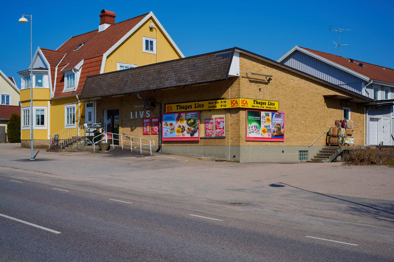 Bilden visar byggnader på en solig dag, med en väg i förgrunden. Till vänster finns en gul byggnad med rött tegeltak som ser ut att vara bostäder, med ett burspråk och en trappa upp till entrén. Framför den gula byggnaden står en ljuslykta. Till höge