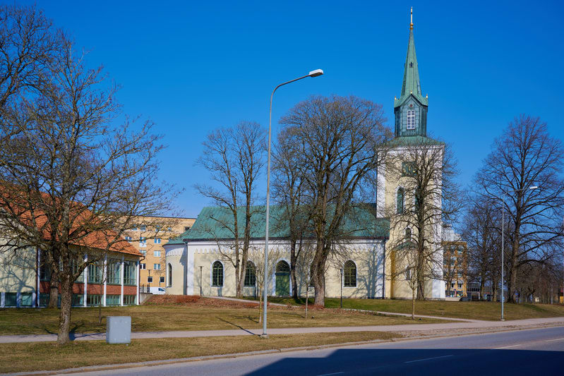 Bilden visar Kyrkan i Vara, en ljusgul kyrkobyggnad med ett grönt tak och en spira, under en klarblå himmel. Framför kyrkan finns en gräsmatta med flera kala träd. Till vänster om kyrkan syns en annan byggnad med tegeltak och stora fönster, och i bak
