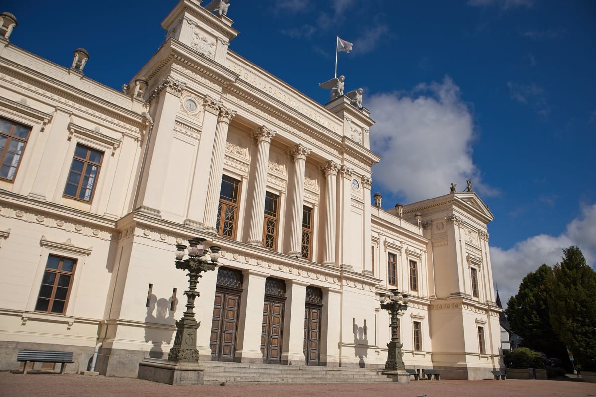 En stor, ljus byggnad i klassisk stil med pilastrar och flera fönster är avbildad under en delvis molnig blå himmel. Byggnaden har detaljerade sniderier och statyer synliga på taklinjen, inklusive statyer av änglar som bär en flaggstång med en vit fl
