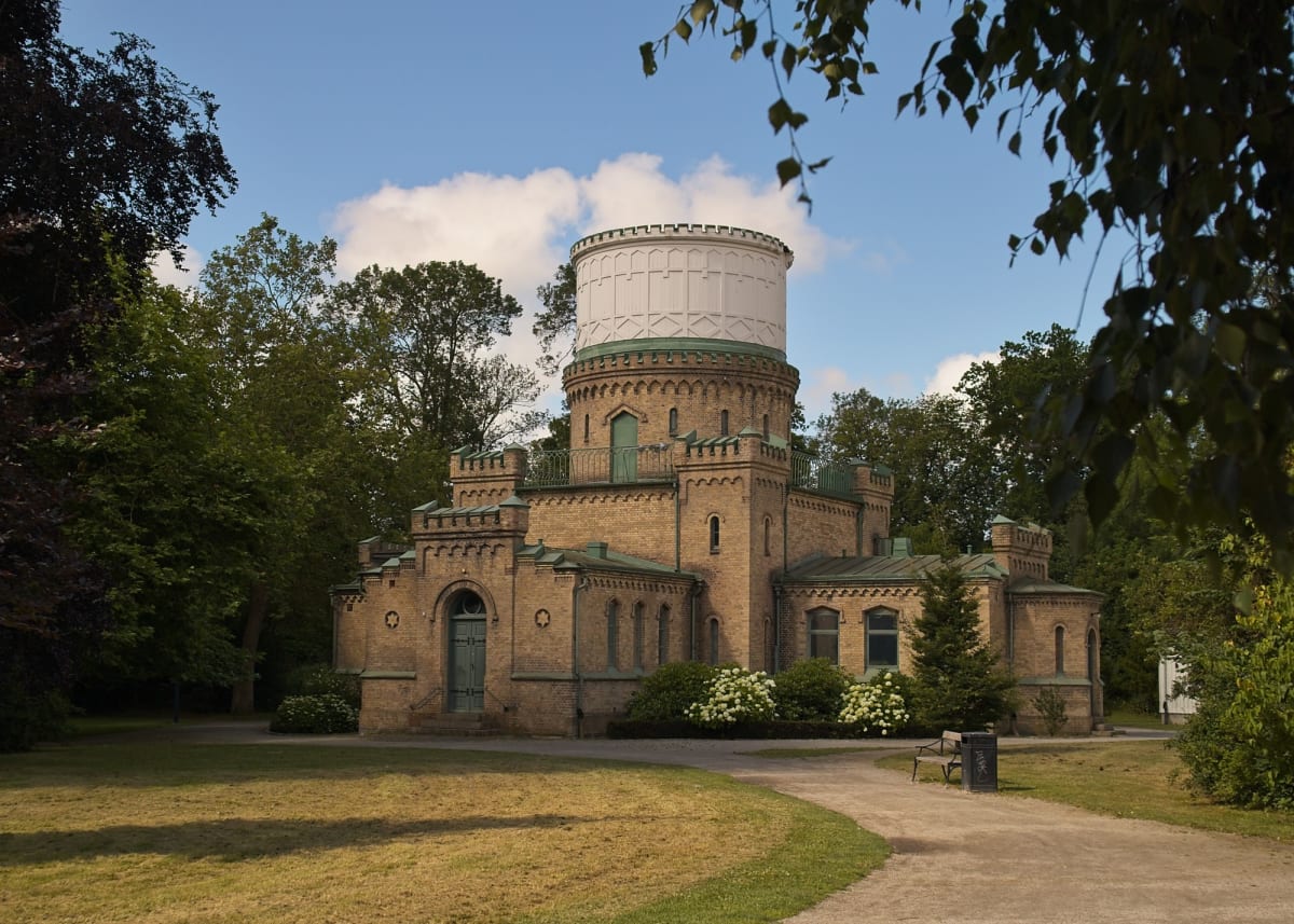 En stor, tegelbyggd byggnad med ett cylindriskt, ljust övre parti, omgiven av grönska under en mestadels blå himmel med några vita moln. Byggnaden har många fönster och dörrar samt en terrass med ett räcke. Arkitekturen inkluderar krenelerade takkant