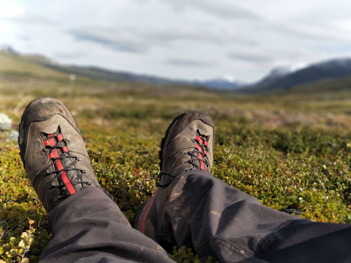Bilden visar ett par fötter iklädda grå vandringskängor med röda detaljer och svarta byxor som vilar på ett gräsbevuxet underlag. Bakgrunden består av ett landskap med kullar och berg, varav några är delvis täckta med snö eller moln. Fjäll. Kungsleden.