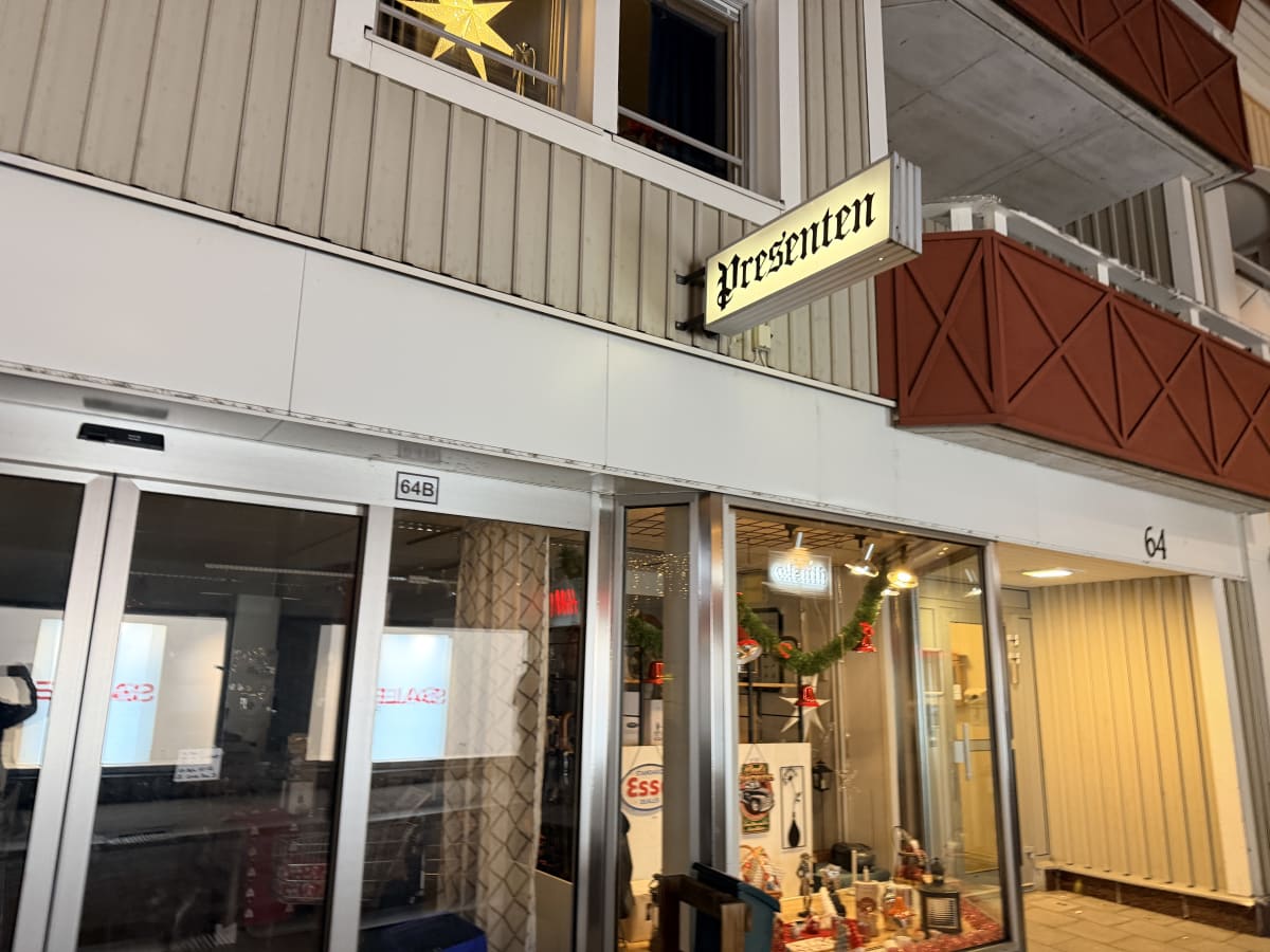 Bilden visar fasaden på en byggnad under kvällstid eller natt. I förgrunden syns en butiksentré med automatiska glasdörrar, märkta '64B', och ett skyltfönster. Över skyltfönstret sitter en rektangulär belyst skylt med texten 'Presenten'. I skyltfönst