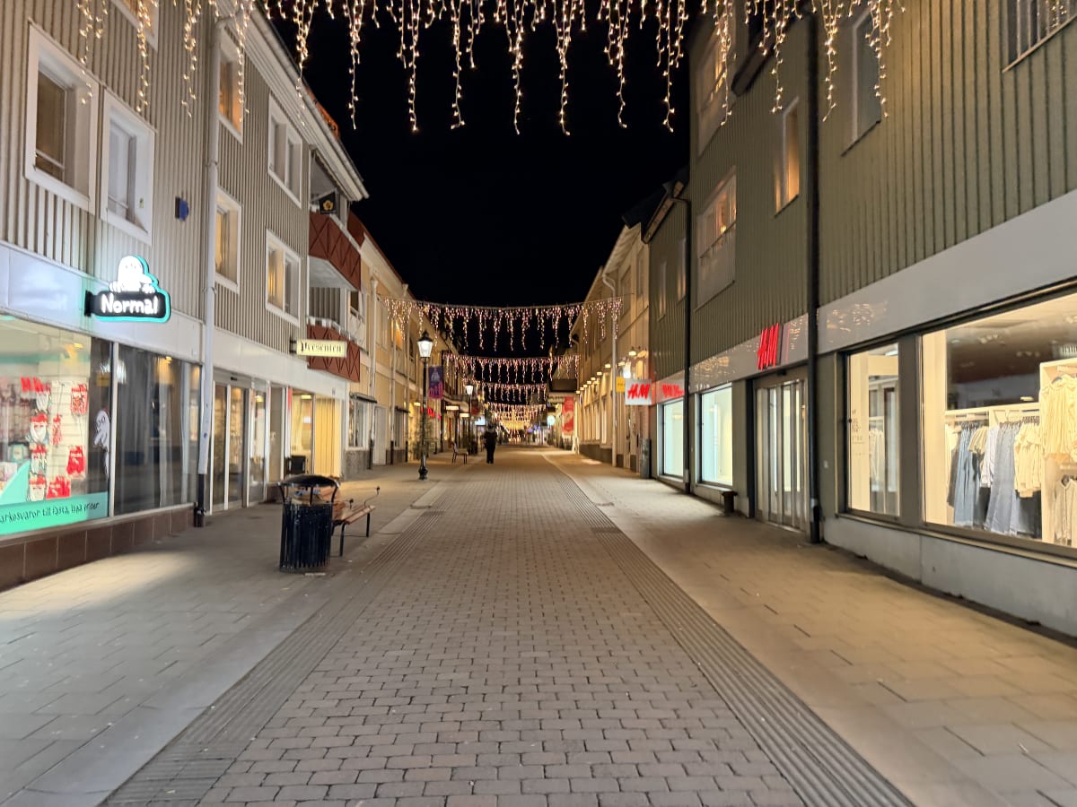 En gata i en stad fotograferad under kvällstid eller natt. Gatan är kantad av byggnader med butiker på bottenvåningarna, identifierbara skyltar syns som "Normal" och "H&M". Över gatan hänger rikligt med julbelysning i form av ljusslingor med droppfor